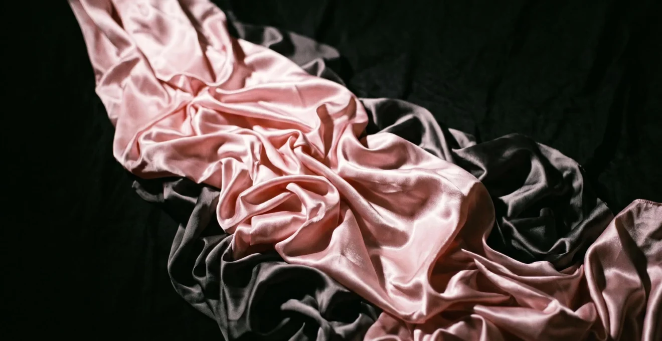 Rouleau de tissu satin rose brillant déroulé de façon chaotique créant plis désordonnés sur fond noir, éclairage dramatique clair-obscur